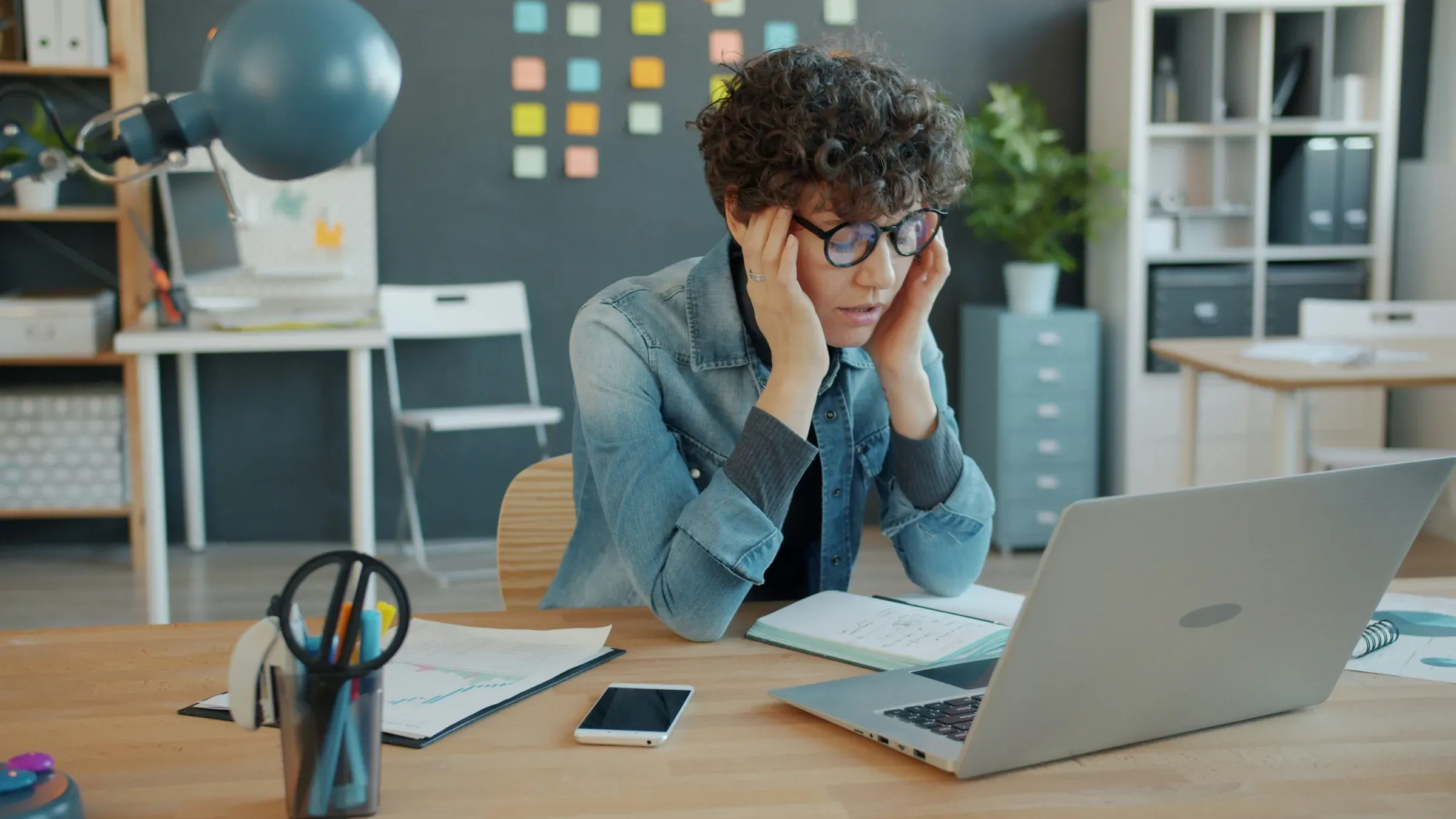 A woman sitting at a cluttered desk with her head in her hands, looking at her laptop