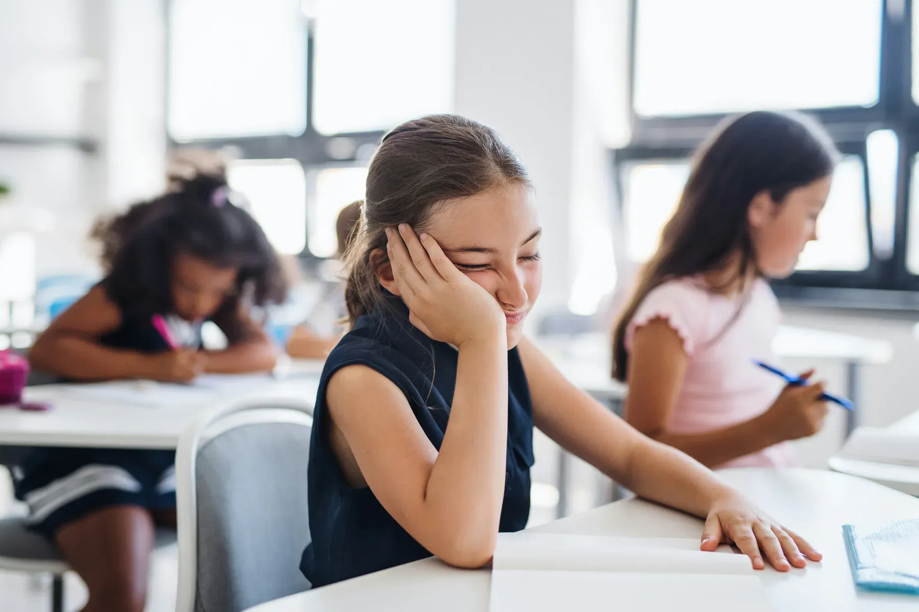 A student resting her head on her hand at a classroom desk while other children work around her