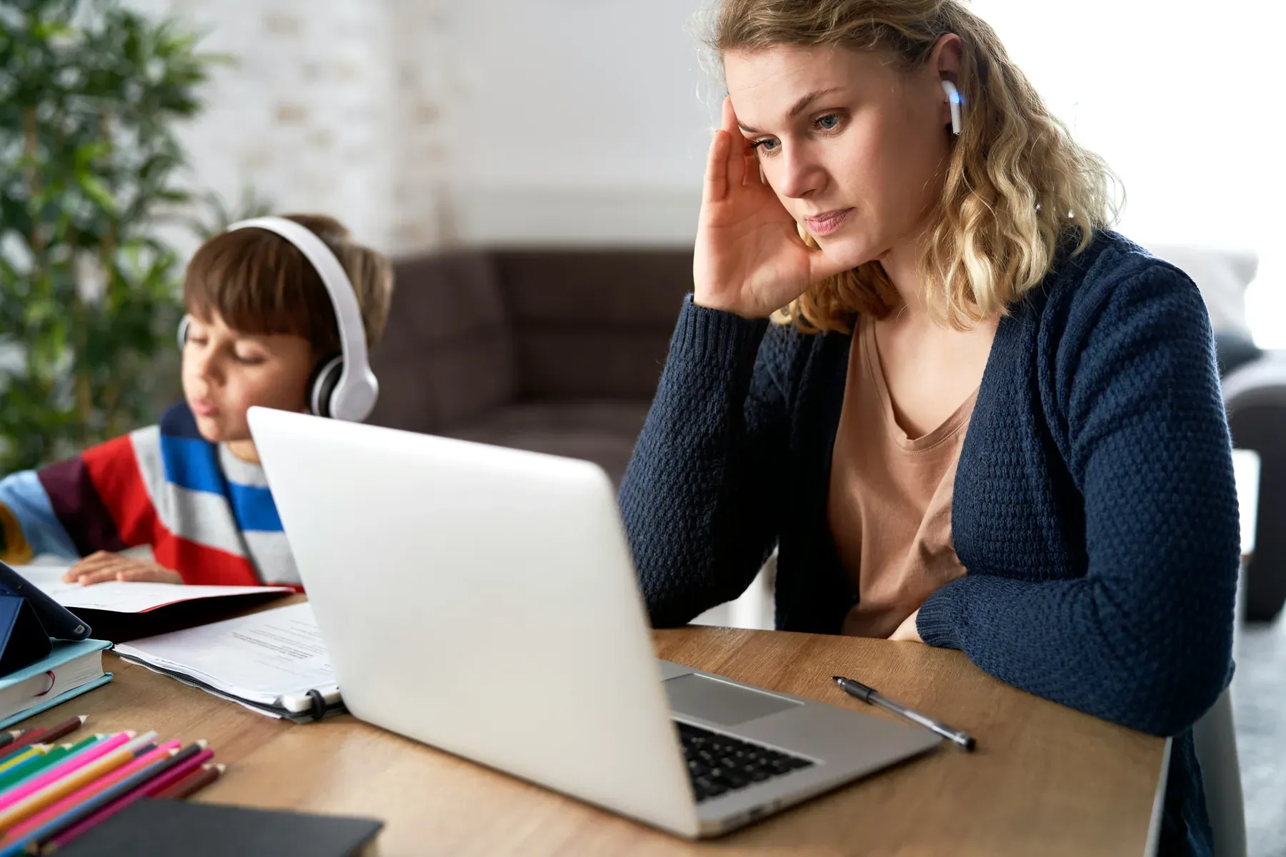 A parent concentrating at a laptop at home while her child works nearby in headphones