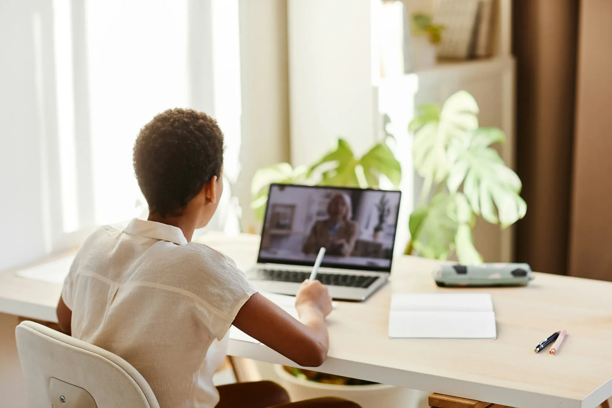 A student writing at a home desk while a woman appears on the laptop screen
