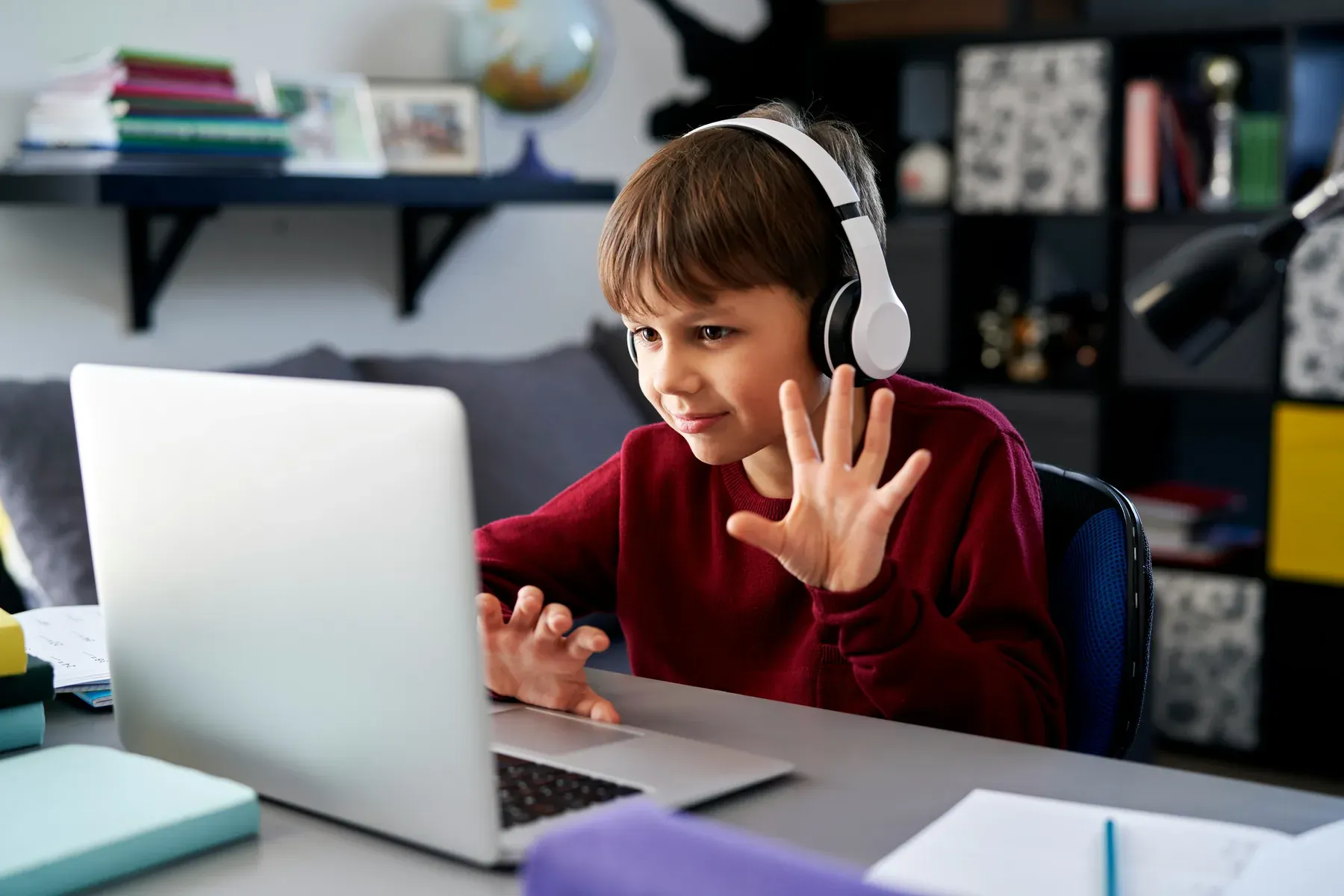 A student in headphones raising a hand to the laptop screen during an online session