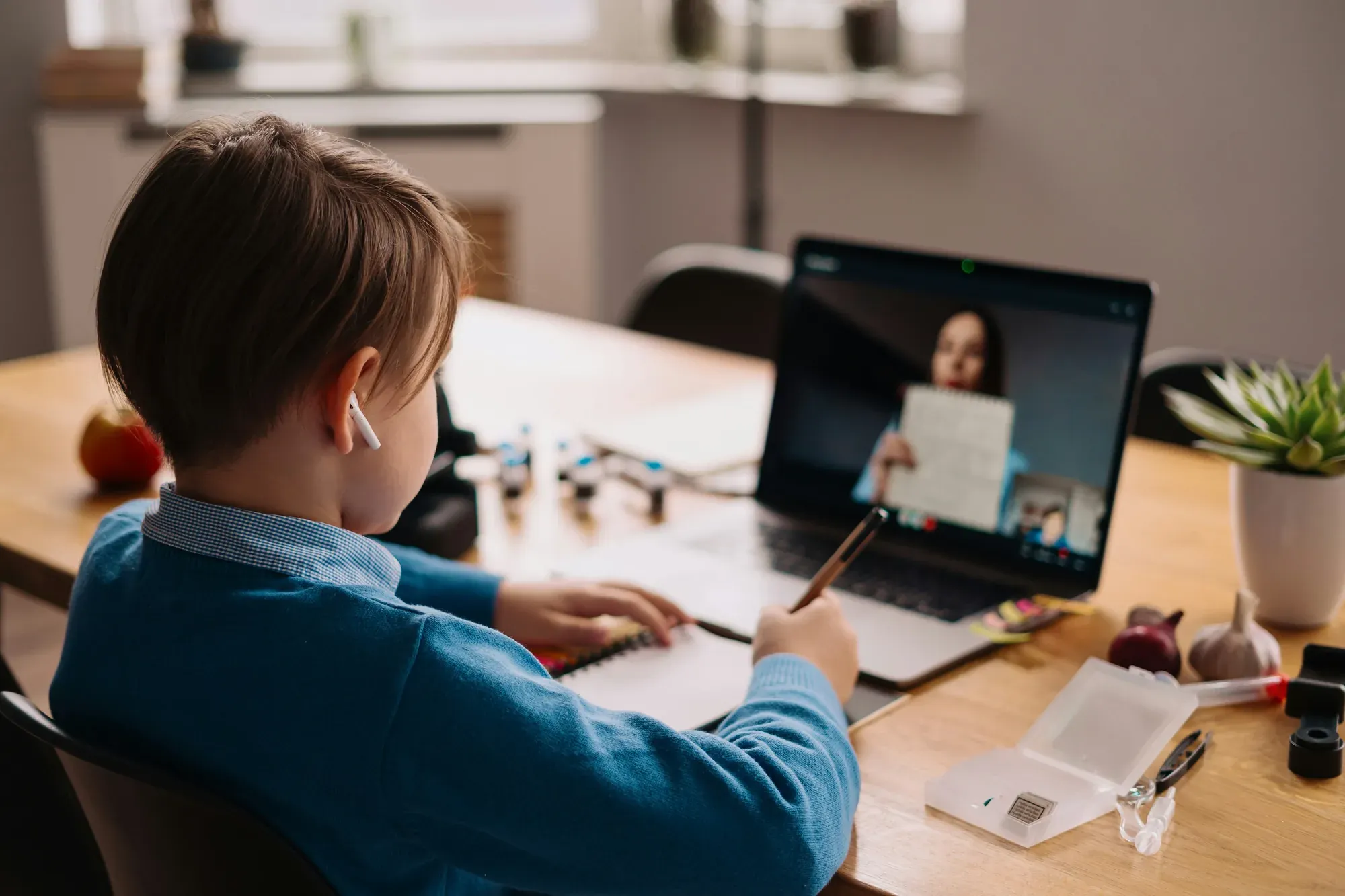 A student writing at a desk while an examiner shows a task on the laptop screen