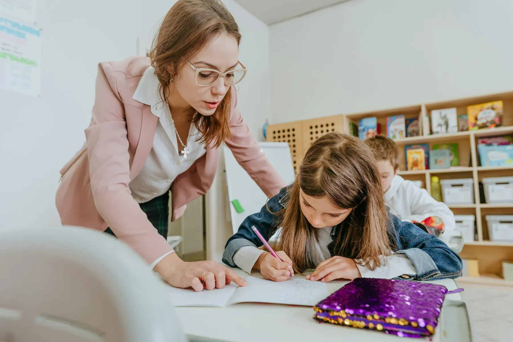 A teacher leaning over a student's desk to help with written work in a classroom